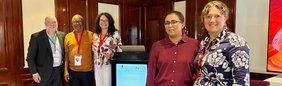 Fünf Teilnehmende eines internationalen Anti-Doping-Seminars stehen neben einem Rednerpult mit Veranstaltungsbanner in einem Konferenzraum.