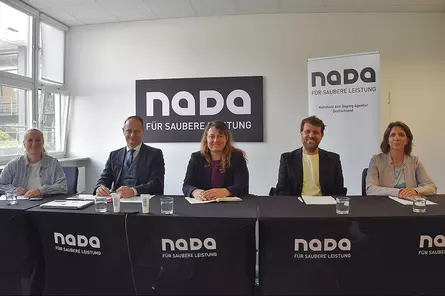 Fünf Personen sitzen an einem Konferenztisch der NADA vor einem NADA-Logo und einem Roll-up-Banner in einem hellen Besprechungsraum.