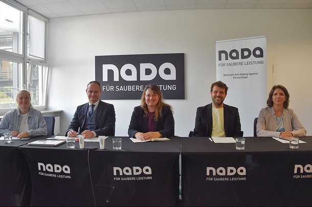 Fünf Personen sitzen an einem Konferenztisch der NADA vor einem NADA-Logo und einem Roll-up-Banner in einem hellen Besprechungsraum.