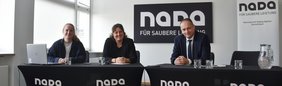 Drei Personen sitzen an Tischen mit NADA-Branding in einem Konferenzraum. Im Hintergrund ist ein NADA-Logo mit dem Schriftzug „Für saubere Leistung“ zu sehen.