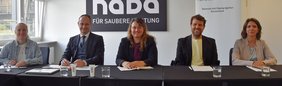 Fünf Personen sitzen an einem Konferenztisch der NADA vor einem NADA-Logo und einem Roll-up-Banner in einem hellen Besprechungsraum.